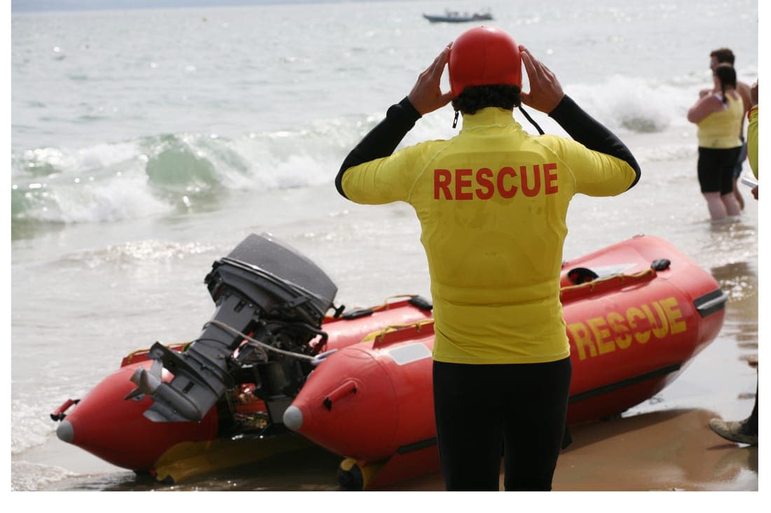 A volunteer lifesaver in a red helmet stands beside an inflatable rescue boat at the water’s edge.